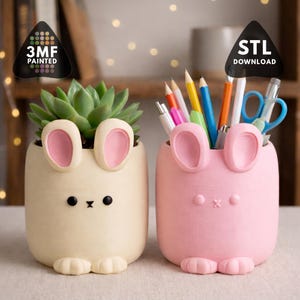 May include: Two bunny-shaped containers, one holding a succulent and the other pencils and pens. The containers are cream and pink with pink inner ears and black eyes. The text "3MF PAINTED" and "STL DOWNLOAD" are visible.