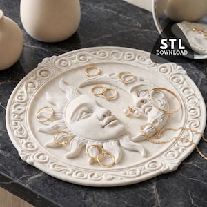 Könnte beinhalten: Ein cremefarbenes, kreisförmiges Schmucktablett mit einem Sonnen- und Monddesign. Das Tablett enthält verschiedene goldene Ringe, ein Armband und eine Halskette. Das Tablett hat einen dekorativen, verzierten Rand und steht auf einer dunklen, strukturierten Oberfläche.