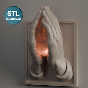 May include: A white wall-mounted sculpture of two hands in a praying position, cradling a small candle with a lit flame. The sculpture is set within a rectangular frame. The text "STL DOWNLOAD" is in a blue circle in the upper left corner.