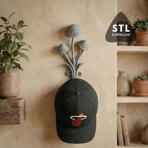 May include: A grey wall-mounted hook shaped like three mushrooms with a black baseball cap featuring a red and white flame logo hanging from it. The text STL DOWNLOAD is visible in the upper right corner of the image, indicating a digital file.