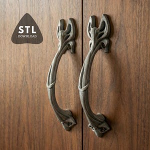Puede incluir: Dos tiradores de armario grises y ornamentados con un diseño curvo, montados en un armario de madera. Los tiradores tienen un patrón detallado de inspiración floral. La imagen también incluye un logotipo que dice "STL DOWNLOAD".