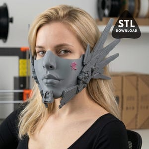 May include: A gray face mask with a stylized design, featuring a nose, lips, and cheekbones. The mask has angular ear pieces and a pink Japanese character on the cheek. The image includes the text "STL DOWNLOAD".