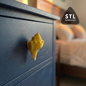 Peut inclure: Un bouton de tiroir en forme de coquillage doré, monté sur une commode bleu marine. Le bouton est un design détaillé en trois dimensions. L'image comprend le texte "STL DOWNLOAD".