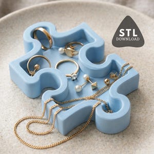 Op de afbeelding: Een lichtblauwe, puzzelvormige sieradenbak met gouden ringen, oorbellen en kettingen. De bak is gemaakt van een stevig materiaal en heeft een matte afwerking. De afbeelding bevat ook een zwarte driehoek met de tekst "STL DOWNLOAD".