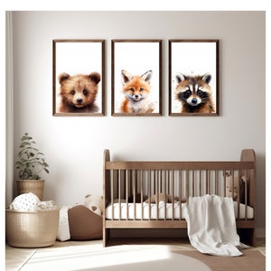 May include: Three framed watercolor paintings of animal faces. The paintings are of a brown bear, a red fox, and a raccoon. The paintings are hung on a white wall above a wooden crib.