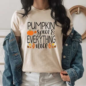 May include: A beige t-shirt with the text "Pumpkin spice & everything nice" in black. A small orange pumpkin with a green stem is above the text. There are small orange and yellow leaf designs around the text.