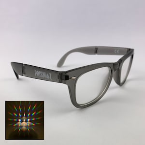 Op de afbeelding: Grijs getinte bril met heldere glazen. De monturen hebben een klassiek ontwerp met de tekst "PRISMAZ" gedrukt op de linker poot. Een kleine inset-afbeelding toont het licht van een kaars dat in een kleurenspectrum wordt gebroken.