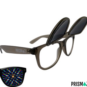 Op de afbeelding: Grijze plastic zonnebril met een omhoogklapbare lensfunctie. De zonnebril heeft een heldere lens en een donkergrijze omhoogklapbare lens. De zonnebril heeft het woord "PRISMAZ" aan de zijkant gedrukt.