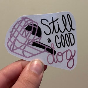 May include: A white sticker with a pink and black illustration of a dog wearing a muzzle. The text "Still a good dog" is written in black cursive.