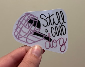 May include: A white sticker with a pink and black illustration of a dog wearing a muzzle. The text "Still a good dog" is written in black cursive.