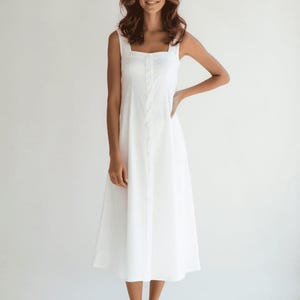 May include: A sleeveless, white midi dress with a square neckline and button-down front. The dress has a fitted bodice and a flared skirt, creating a classic silhouette. The model is wearing the dress.