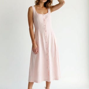 May include: A pale pink, sleeveless midi dress featuring a square neckline and a button-down front. The dress has a fitted bodice and a flared skirt. The model is wearing pink high heels. The dress is made of a lightweight fabric.