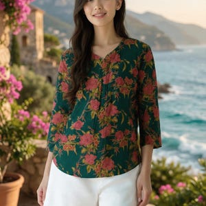 May include: A dark green blouse with a floral pattern of red and yellow flowers. The blouse has a V-neck and three-quarter sleeves. The model is wearing white shorts. The background shows a coastal landscape with mountains and the sea.