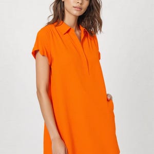 May include: A bright orange short-sleeved shift dress featuring a classic collar, a partial button-down placket, and side pockets. The garment has a straight, relaxed silhouette that falls above the knee, crafted from a solid, smooth fabric.