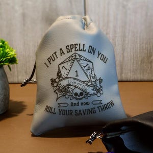 May include: A gray drawstring dice bag with the text "I PUT A SPELL ON YOU" and "ROLL YOUR SAVING THROW". A black dice bag, dice, and a small green plant in a gray pot are also visible. Purple flowers are in the background.