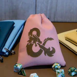 May include: A pink drawstring bag featuring a gold dragon and ampersand logo, alongside multi-coloured dice and matching pouches. The dice are white, blue, and green with gold numbering. The pouches are black, teal, blue, yellow, and grey.