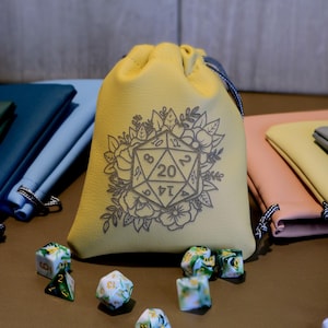 May include: A selection of dice and dice bags in different colours. A yellow dice bag with a floral design and a D20 symbol is central. Other bags are black, green, blue, pink, and grey. Dice are scattered around.