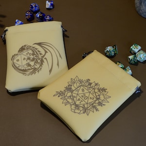 May include: Two beige dice bags with drawstring closures, each featuring a different design. One bag has a dragon and a D20 die, the other has a floral design with a D20 die. Several dice are scattered around the bags.