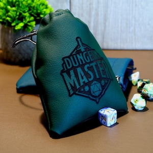 May include: A teal green dice bag with the words "DUNGEON MASTER" printed in black. Several colourful dice are scattered around the bag. The bag is made of a soft material and has a drawstring closure.