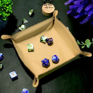Peut inclure: Un plateau de dés en cuir beige aux coins arrondis, contenant plusieurs dés polyédriques colorés. Les dés sont bleus, verts et blancs avec des chiffres dorés. Le plateau est posé sur une surface sombre, avec des plantes décoratives.