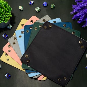 May include: A selection of colourful, square dice trays in shades like black, green, and pink. Each tray features metal eyelets and is designed for tabletop gaming. Dice and decorative plants are scattered around the trays.