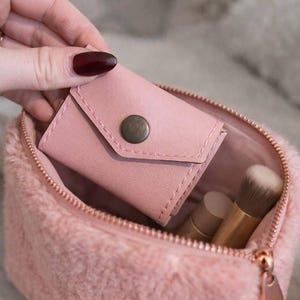 May include: A pink faux fur makeup bag with a rose gold zipper, holding a small pink wallet with a snap closure. Inside the bag are makeup brushes and tubes of makeup. The bag is open, revealing the contents.