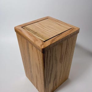 May include: A square wooden waste bin featuring a natural light brown wood grain pattern. The container has a tapered shape with a flat, square lid that sits flush within the top frame, showcasing a smooth finish and visible wood texture.