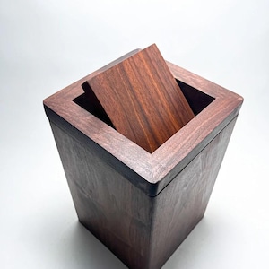 May include: A square dark brown wooden container with a matching lid that is partially open. The box features a visible wood grain pattern throughout its exterior surfaces and a smooth, clean finish on the top edges of the square frame.