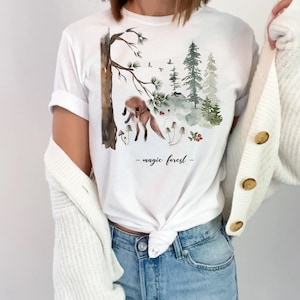 May include: White t-shirt featuring a watercolor illustration of a fox in a forest scene with trees, mushrooms, and berries. The text "magic forest" is printed below the illustration. The shirt is part of a clothing collection.