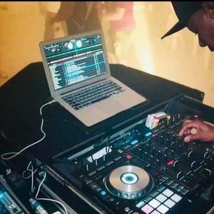 Op de afbeelding: Een zwarte DJ-mixer met een zilveren knop en een laptop met een scherm waarop een muziekprogramma wordt weergegeven. De mixer staat op een zwart oppervlak en een hand ligt op de mixer.