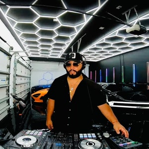 Puede incluir: Un hombre con una camisa negra y una gorra de béisbol negra con un collar de cadena de oro está de pie frente a una mesa de mezclas de DJ. La mesa de mezclas tiene dos tornamesas y un panel de control. El hombre está en un garaje con un techo hexagonal blanco y negro y una pared negra con luces de colores. Hay dos coches en el fondo.