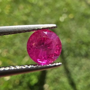 Puede incluir: Una gema redonda, facetada, de color rosa intenso, sostenida por unas pinzas plateadas. La gema es de un color magenta vibrante, con estructuras internas visibles y reflejos de luz. El fondo es verde borroso.