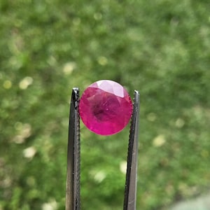 Pode incluir: Um rubi redondo e facetado, segurado por uma pinça. O rubi é de um vermelho vibrante com um ligeiro tom rosado. O fundo é verde desfocado, sugerindo um ambiente ao ar livre. A pedra preciosa é o ponto focal.