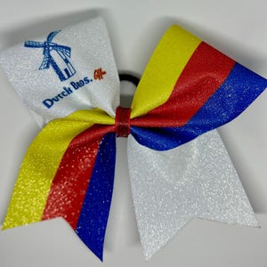 May include: A glittery cheer bow with a white section featuring the text "Dutch Bros." and a windmill logo. The bow has yellow, red, and blue stripes. The bow is tied with a red center.