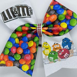 May include: A glittery cheer bow featuring a colorful M&M's candy design. The bow has a white base with the M&M's logo and cartoon characters. The bow is decorated with red, yellow, blue, green, and orange candies.