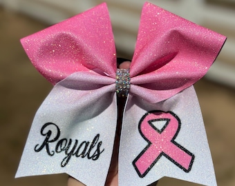 Peut inclure: Noeud de cheerleader en paillettes rose et blanc avec un ruban noir et le mot "Royals" d'un côté et un ruban rose avec un contour noir de l'autre.