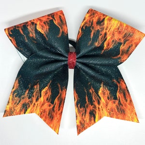May include: A large, glittery black hair bow with a fiery orange and yellow flame design. The bow is tied with a red glitter center and has a black elastic band. This accessory is perfect for cheerleading or adding a bold touch to any hairstyle.