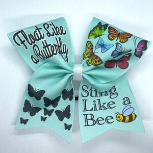 May include: A light blue cheer bow with the text "Float Like a Butterfly" on one side and "Sting Like a Bee" with a cartoon bee on the other side. The bow is decorated with black butterfly silhouettes and colorful butterfly illustrations.