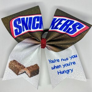 May include: A glittery hair bow featuring a Snickers candy bar theme. The bow has brown and white sections with the Snickers logo in blue and red. The text "You're not you when you're Hungry" is also visible.