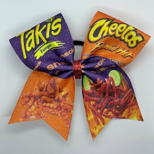 May include: A purple and orange cheer bow with a Takis Fuego and Cheetos Flamin' Hot design. The bow is decorated with glitter and has a black elastic band.