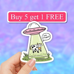 May include: A sticker featuring a cartoon cow being abducted by a UFO. The UFO is pink and green and has the text "Ooh, Shinny!" The sticker also has the text "Buy 5 get 1 FREE".