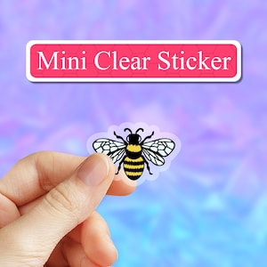 May include: A clear sticker with a black and yellow bee illustration. The sticker is being held by a hand against a pink and blue background. The text "Mini Clear Sticker" is visible above the sticker.