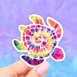 May include: A colorful tie-dye turtle sticker with a white outline. The turtle is facing the right side of the image. The sticker is being held by a hand.