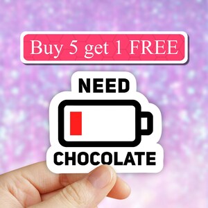 May include: White sticker with black text that reads "NEED CHOCOLATE" and a black and white battery icon with a red bar indicating low battery. The sticker is being held by a hand.