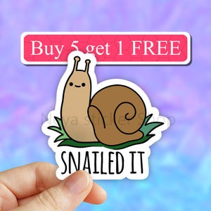May include: A white sticker with a cartoon snail on a green leaf. The snail is brown with a light brown shell. The text "SNAILED IT" is below the snail. The sticker has a pink banner at the top that says "Buy 5 get 1 FREE".