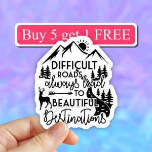 May include: Black and white sticker with a mountain range, a deer, and an arrow pointing to a winding road. The text reads "Difficult roads always lead to beautiful destinations".