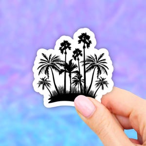 May include: A black and white sticker featuring a tropical island scene. The design showcases several palm trees of varying heights and styles, creating a silhouette against a white border. The sticker is held against a colorful, blurred background.