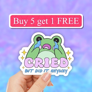May include: A green frog sticker with a pink background and the text "Cried but did it anyway". The frog is crying with sparkling tears. The sticker has a white border and a yellow star on each side.