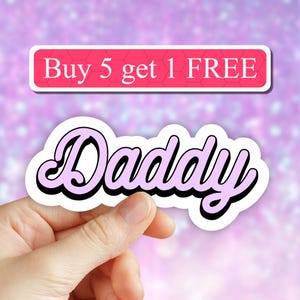 May include: Pink "Daddy" sticker with a white outline and black shadow. The sticker is being held by a hand. The text "Buy 5 get 1 FREE" is visible in the background.