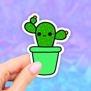 May include: A green cactus sticker with a smiling face and small orange flowers. The cactus is in a green pot with a white border. The sticker is held by a hand with pink nail polish against a purple and blue background.